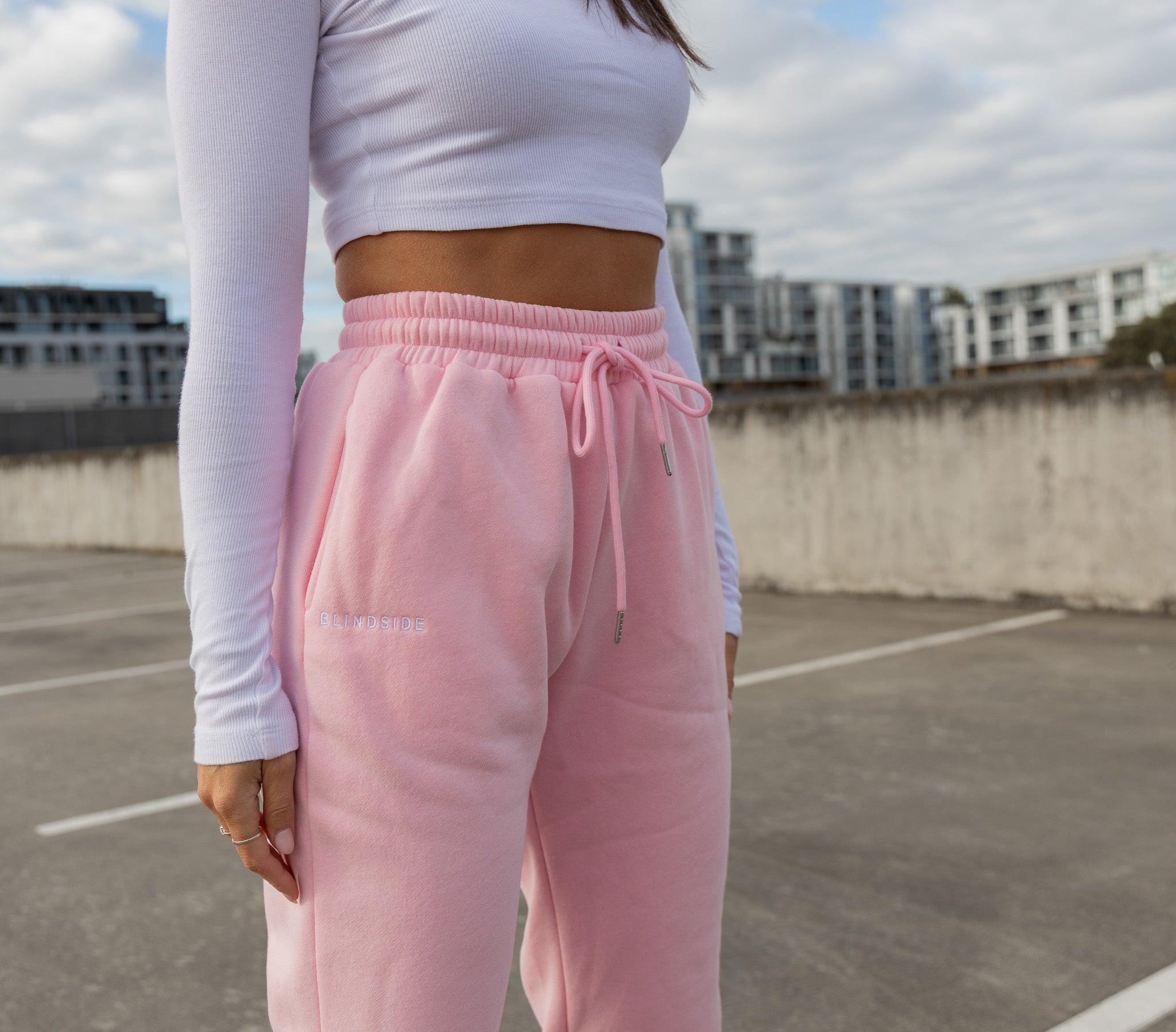 A person in BLINDSIDE’s Society Sweatpants - Pink stands in an outdoor parking lot with buildings behind them. The photo, cropped at the shoulders, shows the pants paired with a white long-sleeve crop top.
