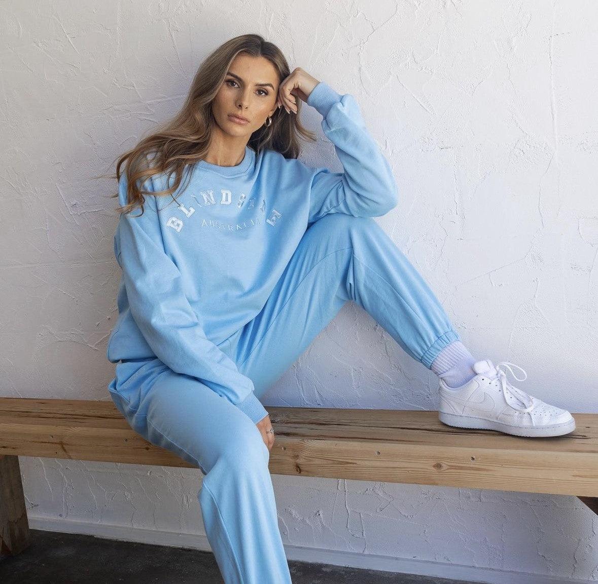 A woman sits on a wooden bench against a white textured wall, wearing BLINDSIDE Legacy Pants in light blue with a matching sweatshirt and white sneakers—one leg bent on the bench, the other foot resting on the floor.