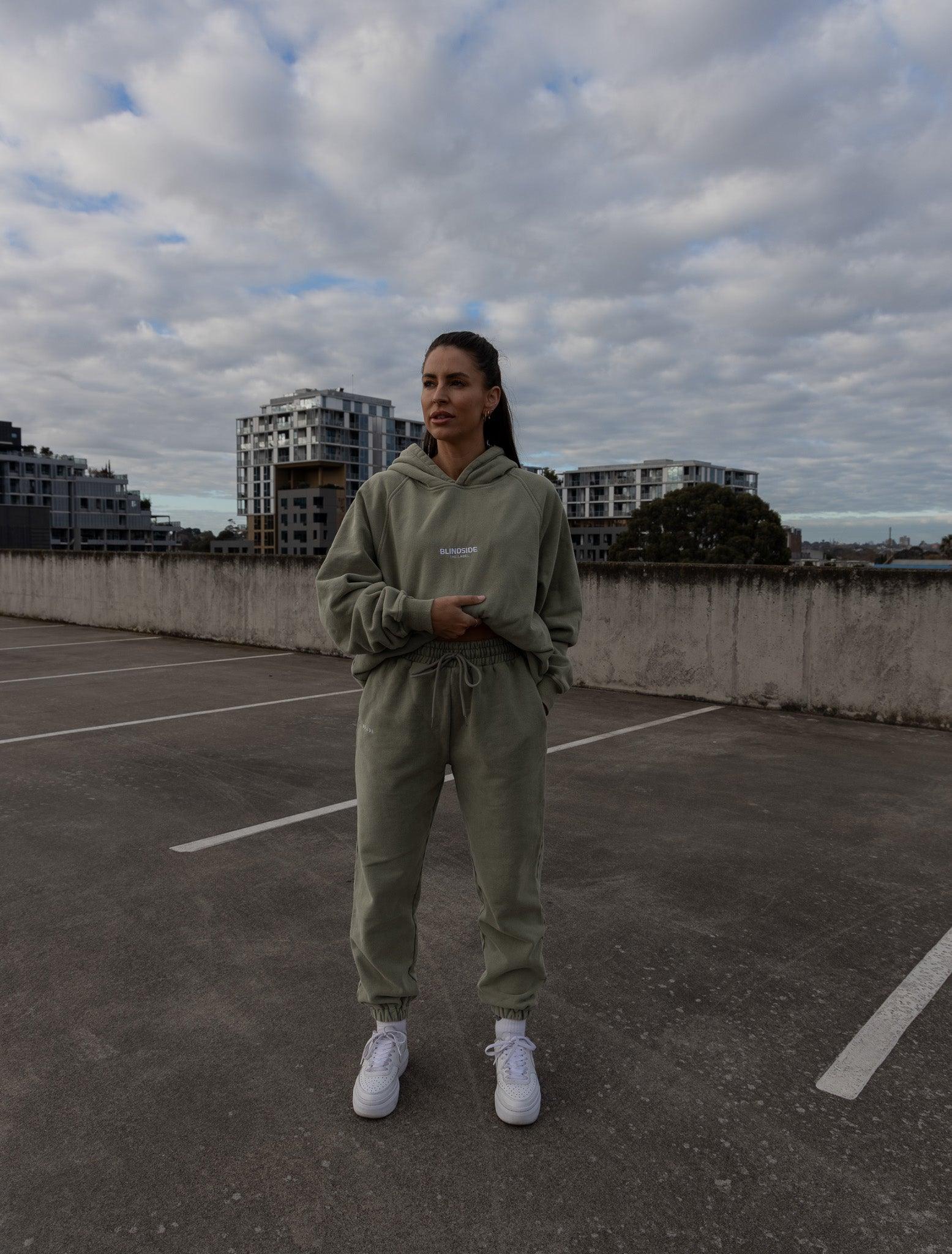 A person stands on a rooftop in BLINDSIDEs Mid Town Sweatpants - Olive, made from cozy 470 GSM fleece. Paired with an olive hoodie and white sneakers, this unisex look is set against modern buildings and an overcast sky.