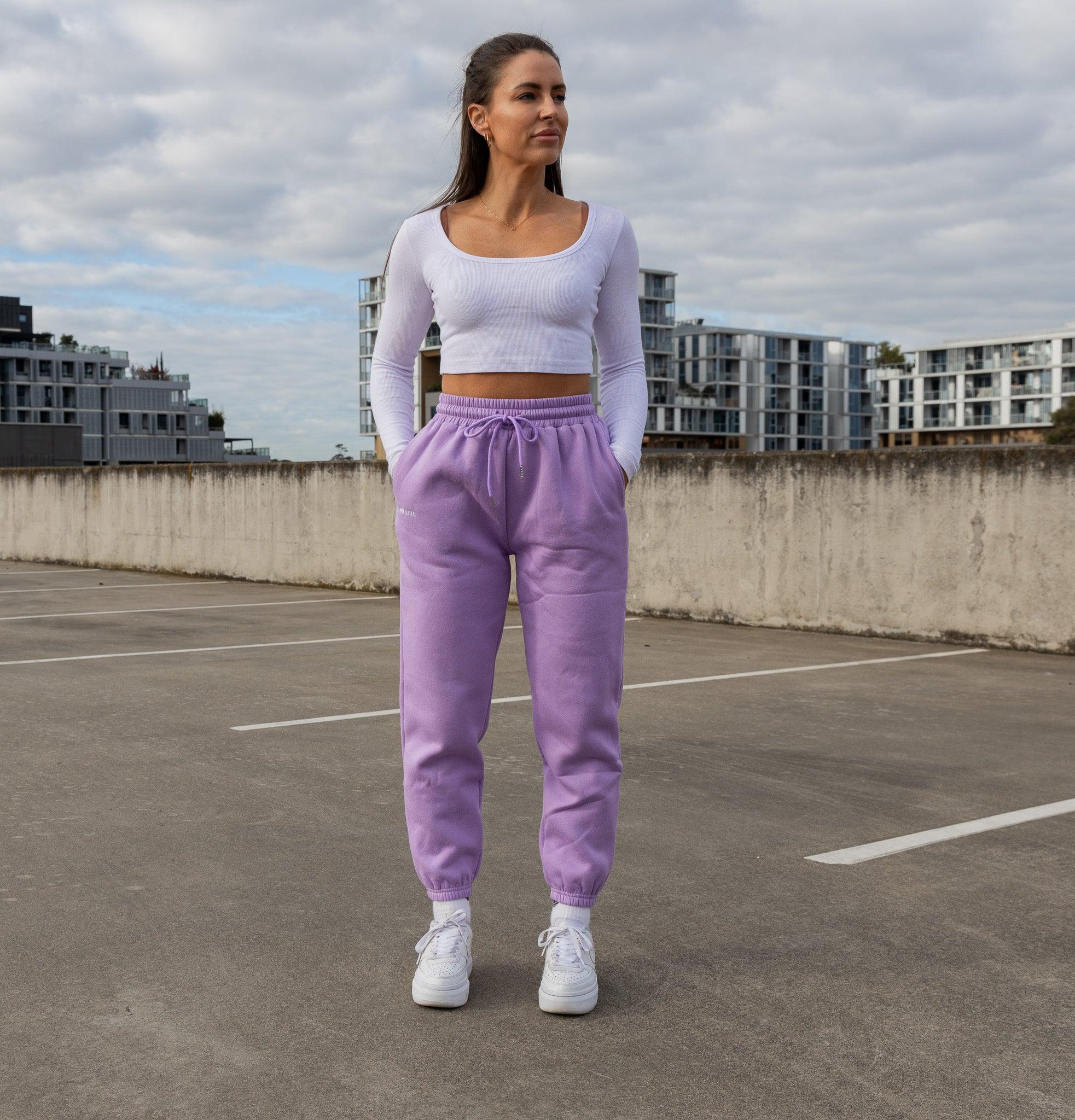 A woman stands in an empty parking lot wearing BLINDSIDE’s Society Sweatpants in Lilac, a long-sleeve white crop top, and white sneakers. She has her hands in her pockets and looks off to the side with apartment buildings behind her.