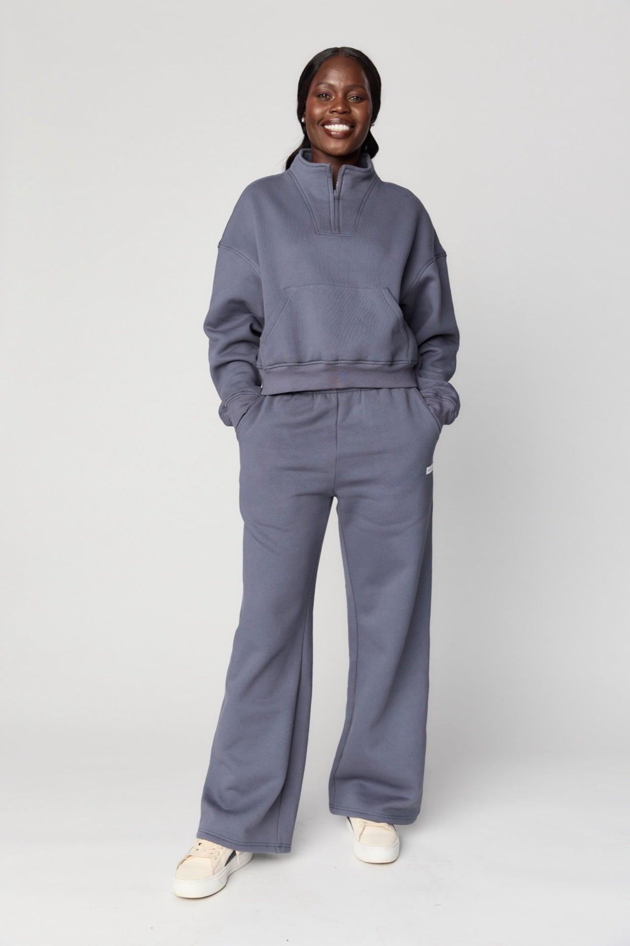 A person smiles with hands in pockets, wearing the BLINDSIDE Main Street 1/4 Zip Jumper in Faded Navy against a plain background, paired with matching sweatpants and white sneakers.