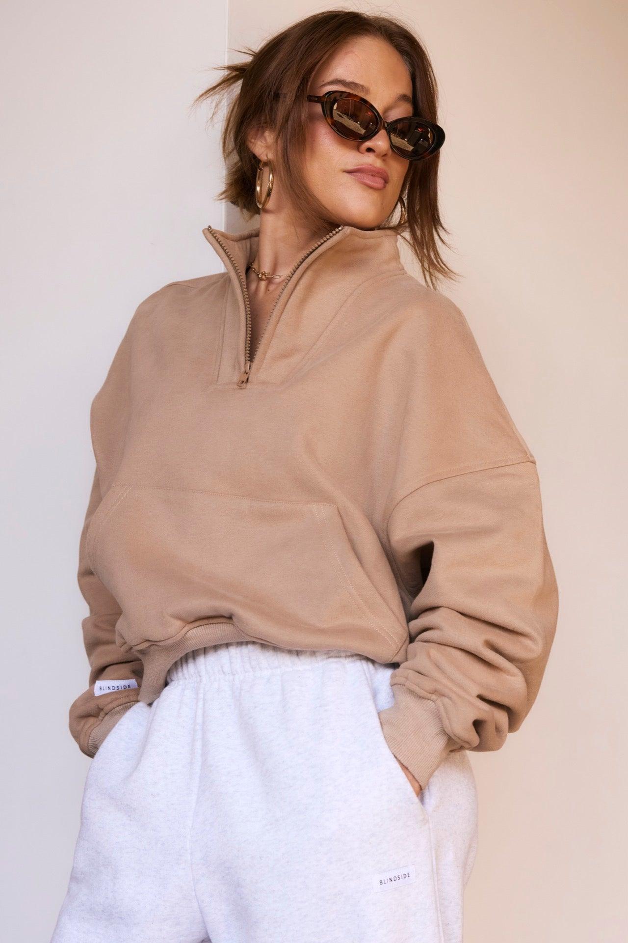 A woman models the BLINDSIDE Main Street - 1/4 Zip Jumper in Latte, paired with light gray sweatpants, sunglasses, and hoop earrings; her hair is tied back with loose strands framing her face against a plain background.