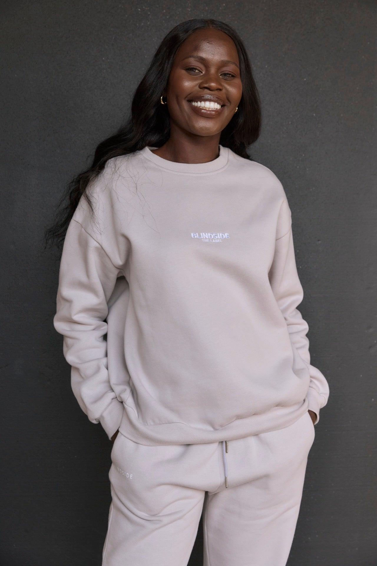 A person stands against a dark background, smiling and wearing BLINDSIDEs Home State Sweatshirt in Light Stone—an oversized, premium cotton blend piece with hands tucked into the pockets.