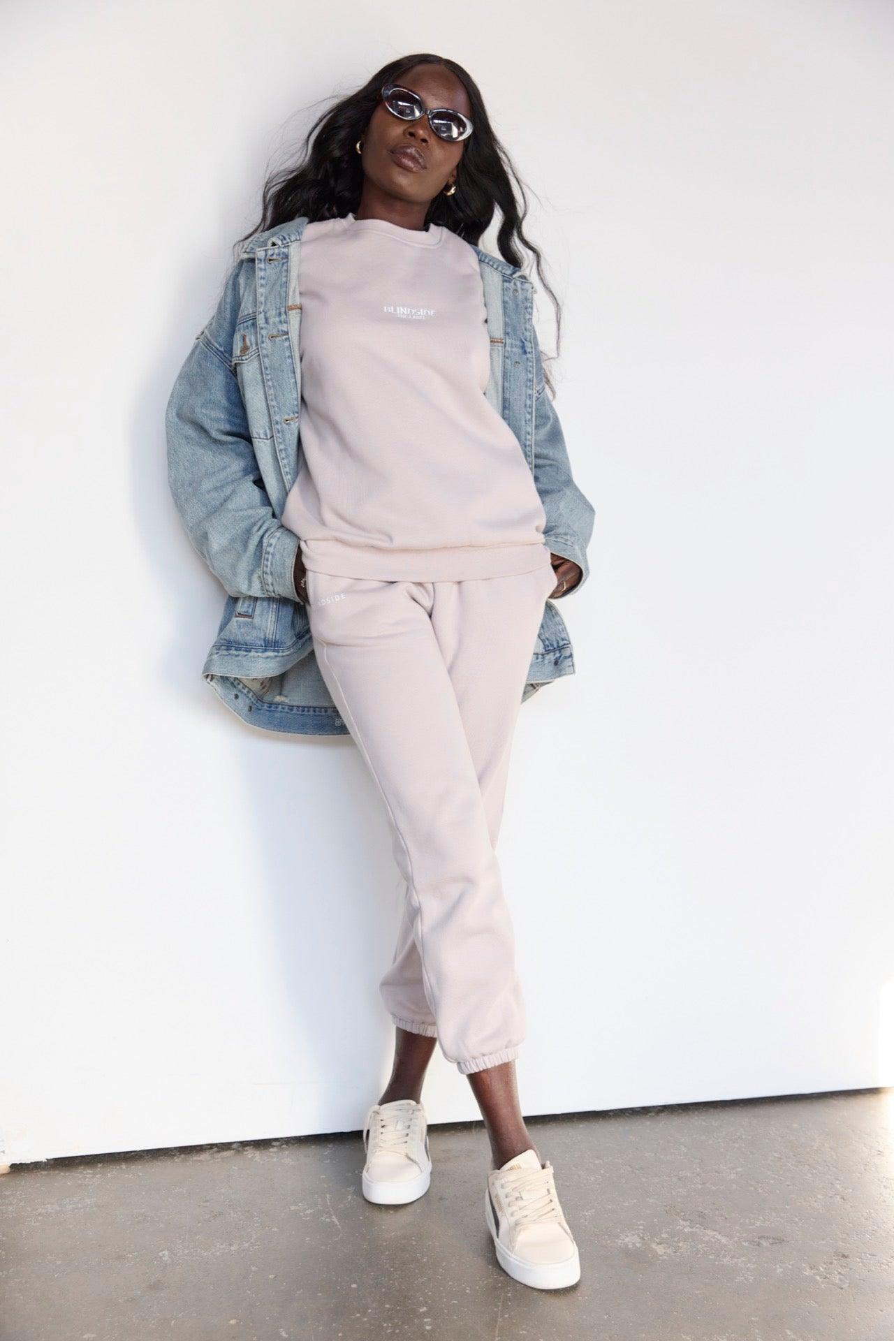 A person stands against a white wall wearing BLINDSIDE’s Home State Sweatpants in Light Stone with an elastic waistband and deep pockets, styled with a light pink sweatshirt, blue denim jacket, sunglasses, and beige sneakers. One leg is slightly bent.