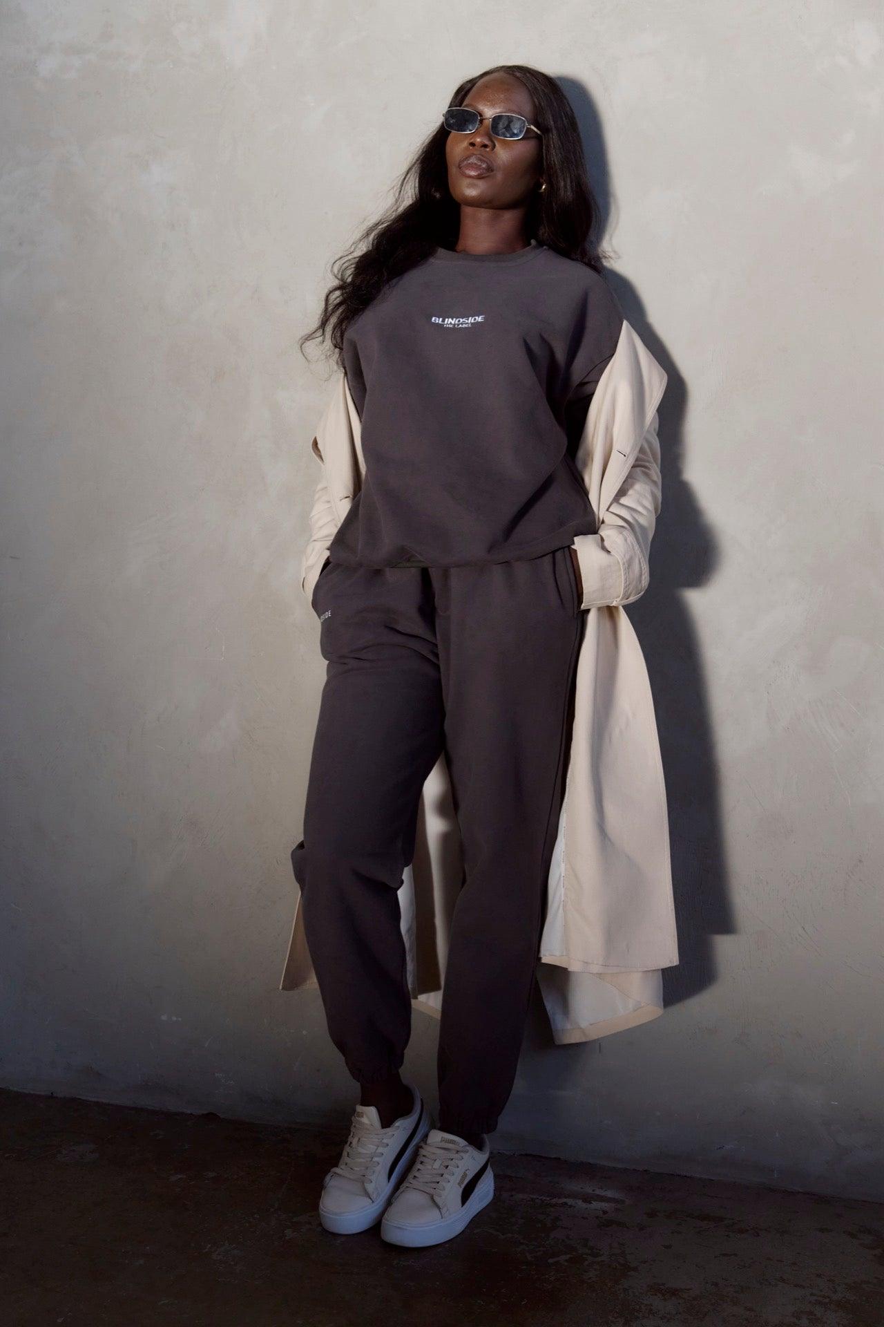 A woman stands against a light gray wall, hands in deep pockets, wearing BLINDSIDEs Home State Sweatpants in Charcoal with an adjustable waistband, alongside a dark sweatshirt, light trench coat, sunglasses, and white sneakers with black accents.