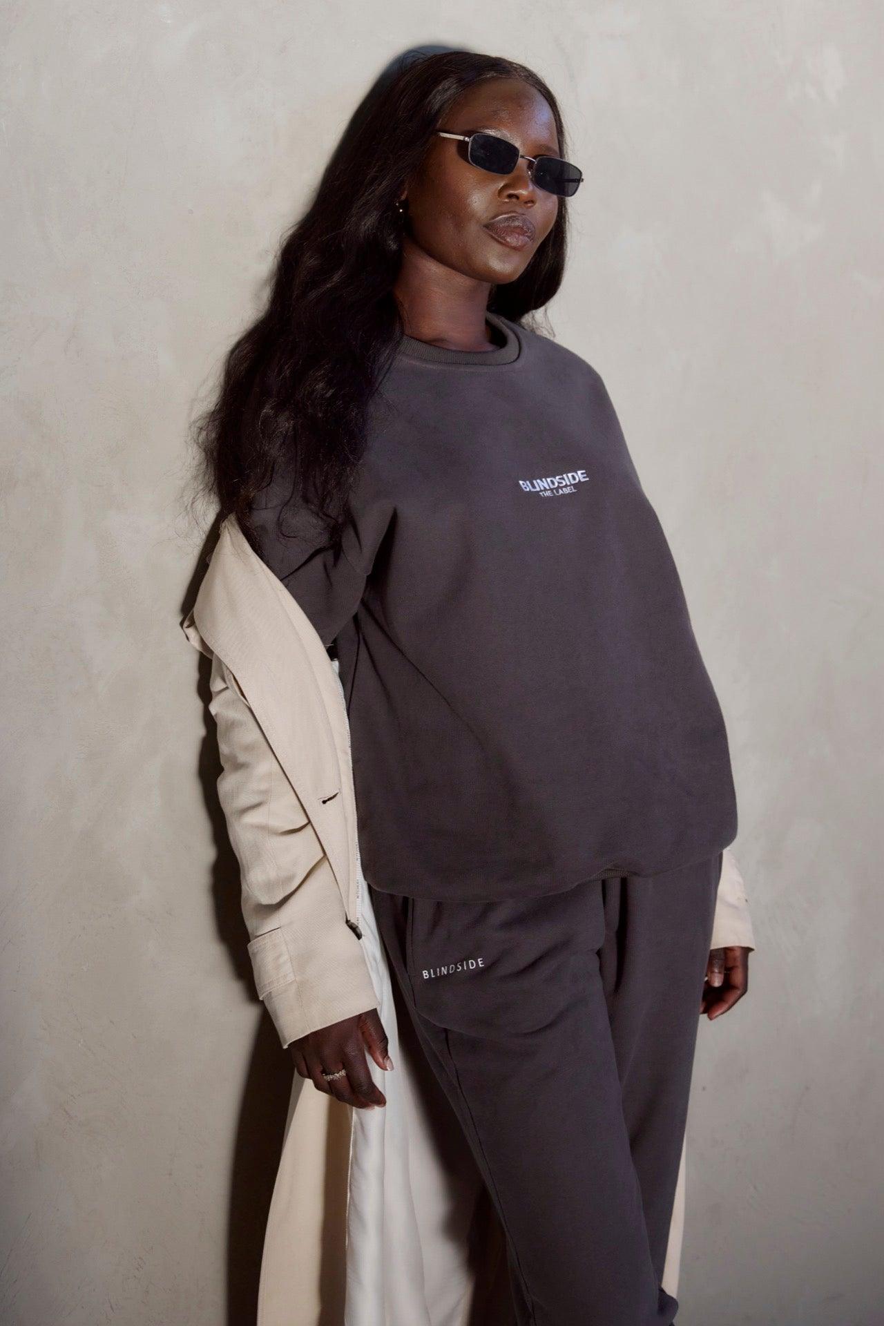 A person with long dark hair wears sunglasses and the BLINDSIDE Home State - Sweatshirt - Charcoal with matching sweatpants, plus a light coat over their shoulders, standing against a plain light wall.